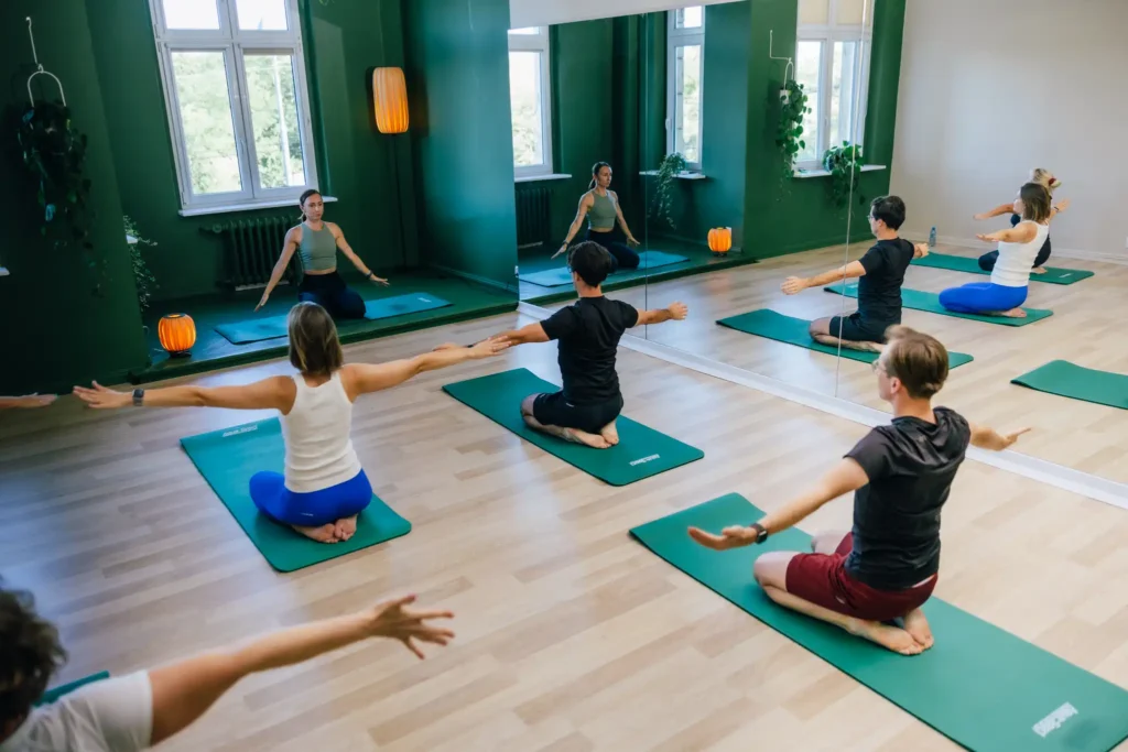 Grupa ludzi, głównie kobiet, siedzących na matach do jogi i wykonujących pozycje jogi w studiu z zielonymi ścianami